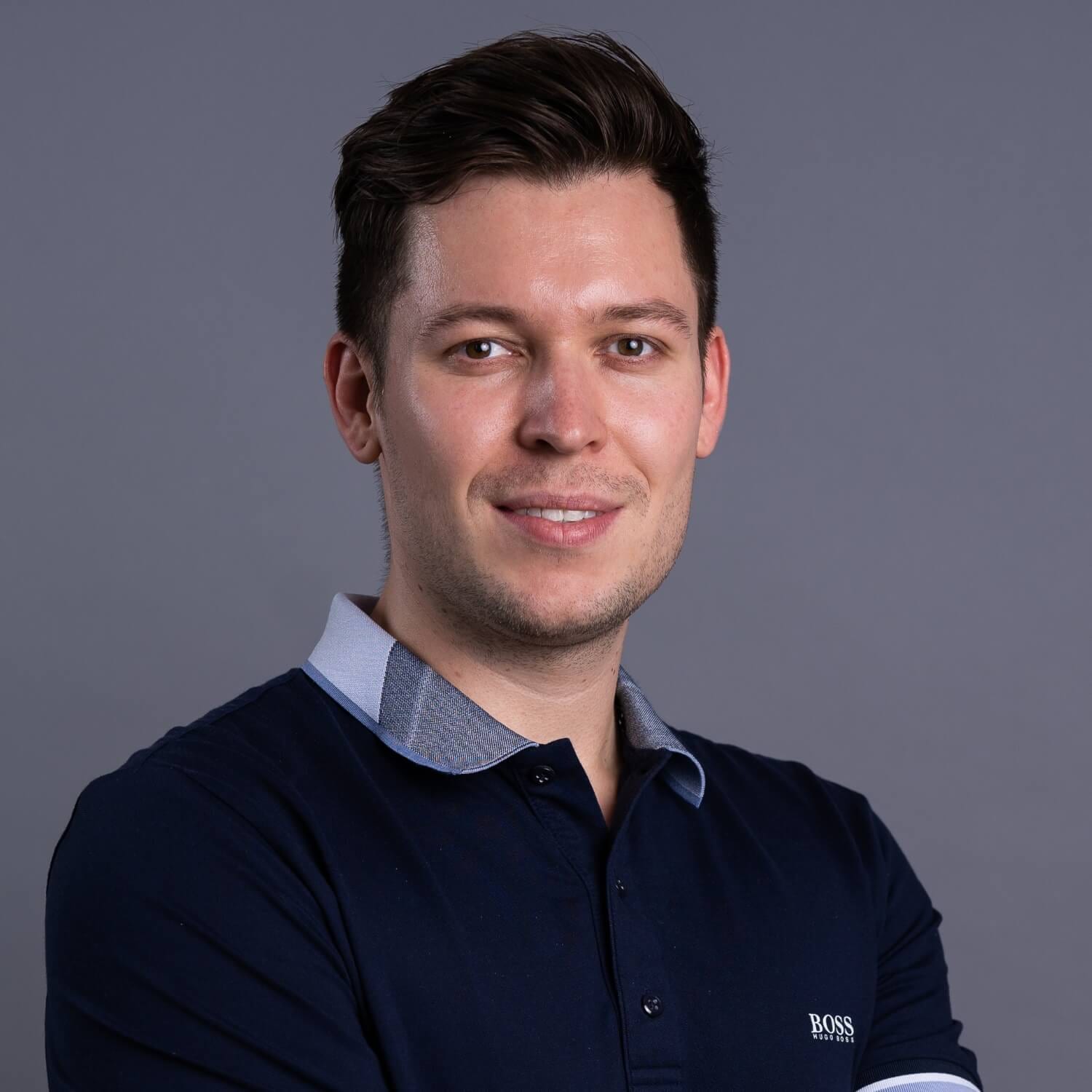 Studioaufnahme: Mann mit dunklem Polohemd, lächelnd, vor neutral grauem Hintergrund.