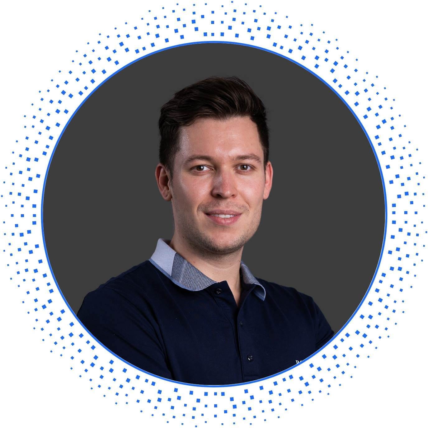 Porträt eines Mannes mit dunklem Haar und Poloshirt vor grauem Hintergrund, freundlich lächelnd.