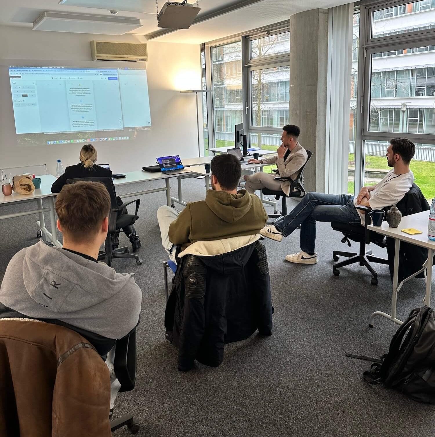 Teilnehmende sitzen in einem Seminarraum und verfolgen eine Präsentation am Beamer; Laptops und Notizen auf den Tischen.