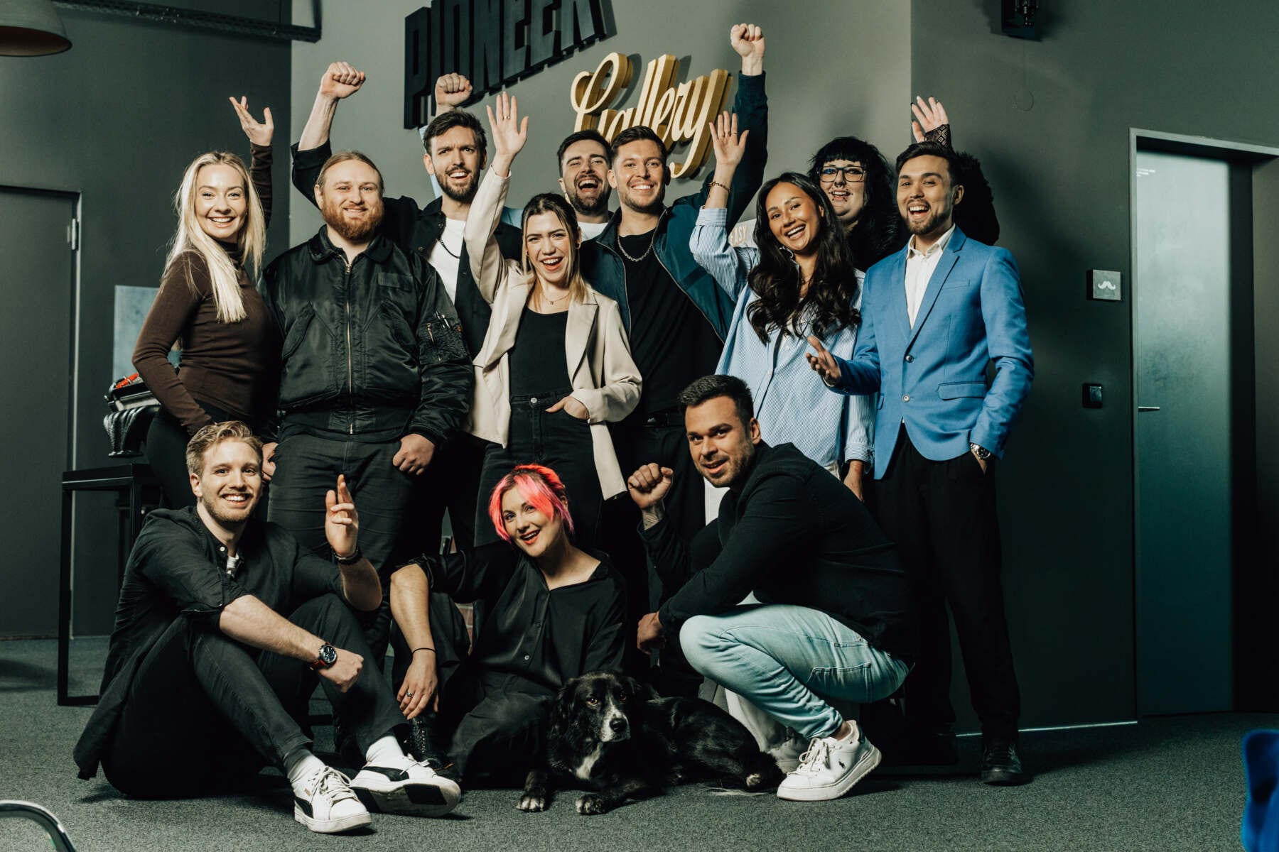 Gruppenfoto eines Teams im Büro, mehrere Personen jubeln und lächeln, ein Hund liegt vorne auf dem Boden.